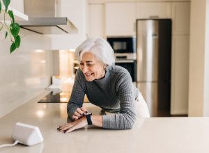 Senior woman smiling happily while using smart devices in her kitchen. Cheerful elderly woman using a home assistant to perform tasks at home.